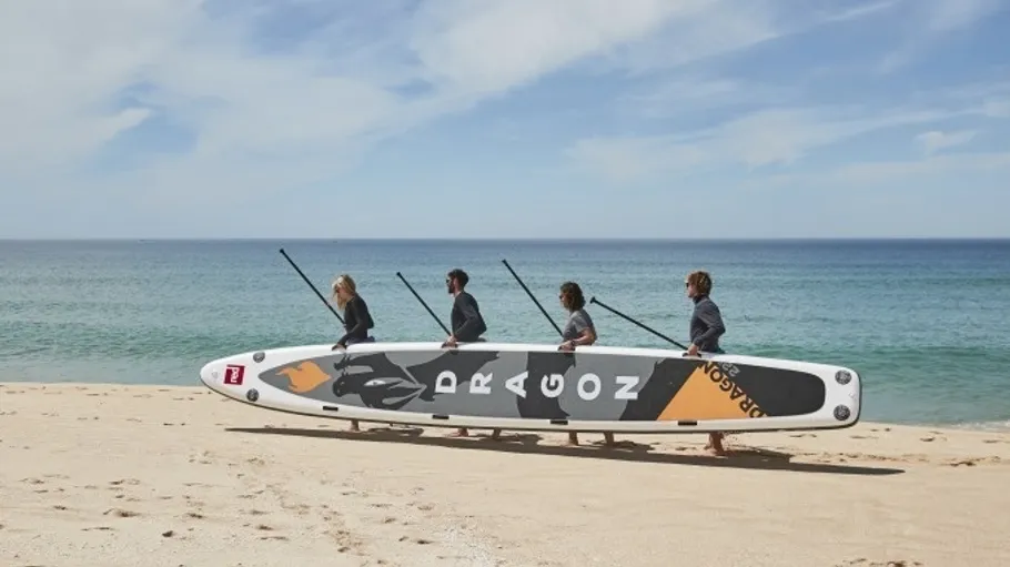 Vier Personen tragen Paddleboard am Strand.