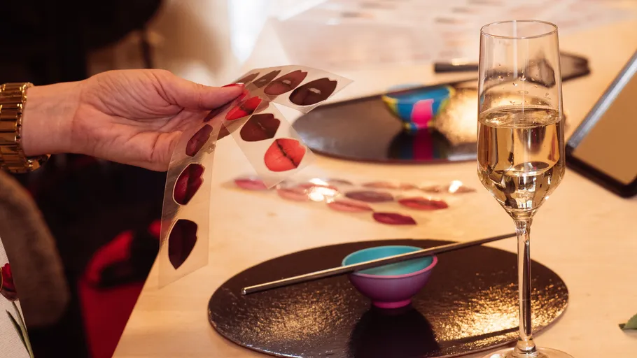 Hand hält Lippenstiftmuster neben Champagnerglas.