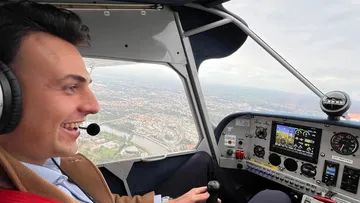 Pilot smiling, flying small plane, city below.