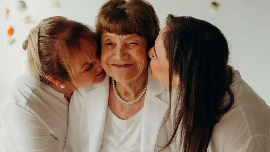 Two women kiss elderly woman indoors.