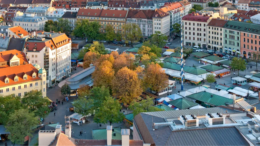 Marktplatz mit Ständen und Bäumen, umgeben von Gebäuden.