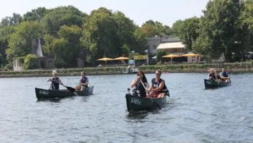 People paddling canoes on a lake, trees in background.