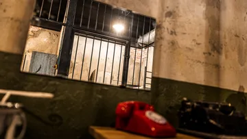 Red phone, desk, barred window, dim room.