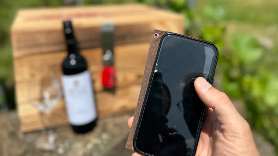 Hand hält Handy neben Weinflasche draußen.