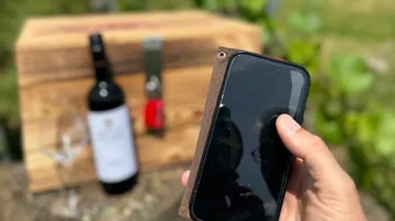 Hand holding smartphone near wine bottle outdoors.