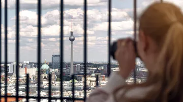 Woman photographing cityscape through bars, Berlin skyline visible.