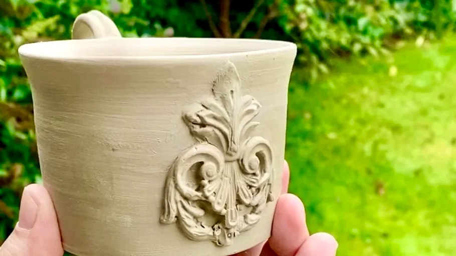 Hand holding decorated clay cup, greenery background.
