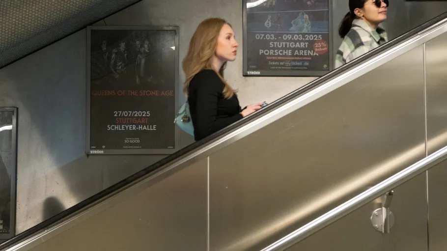 Zwei Frauen auf Rolltreppe, Konzertplakate dahinter.