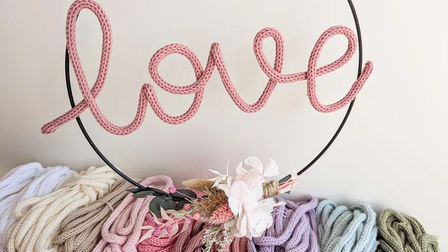 Pink yarn spells 'love' on round frame, surrounded by colored yarn.