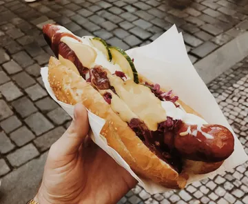 Hand holding hot dog with toppings outdoors.