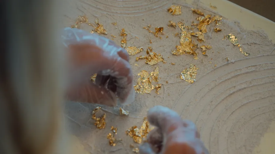 Hands applying gold leaf to textured surface.