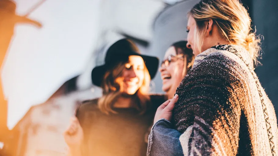 Three women laughing outdoors in sunlight.