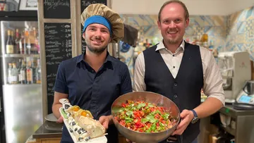 Two men smiling, holding dishes in a restaurant.