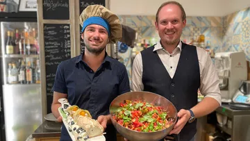 Two smiling men holding food plates, kitchen background.