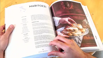 Hands holding cookbook open to Maritozzi recipe.