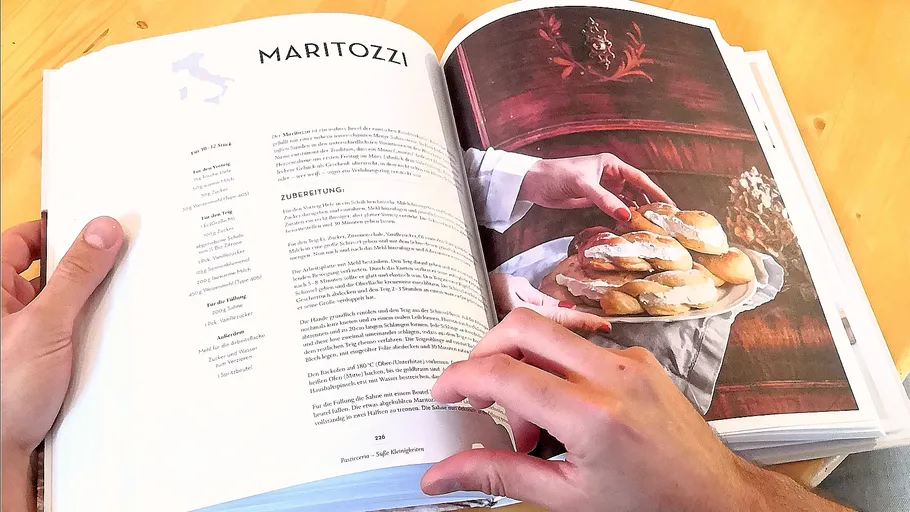 Hands holding cookbook open to Maritozzi recipe.