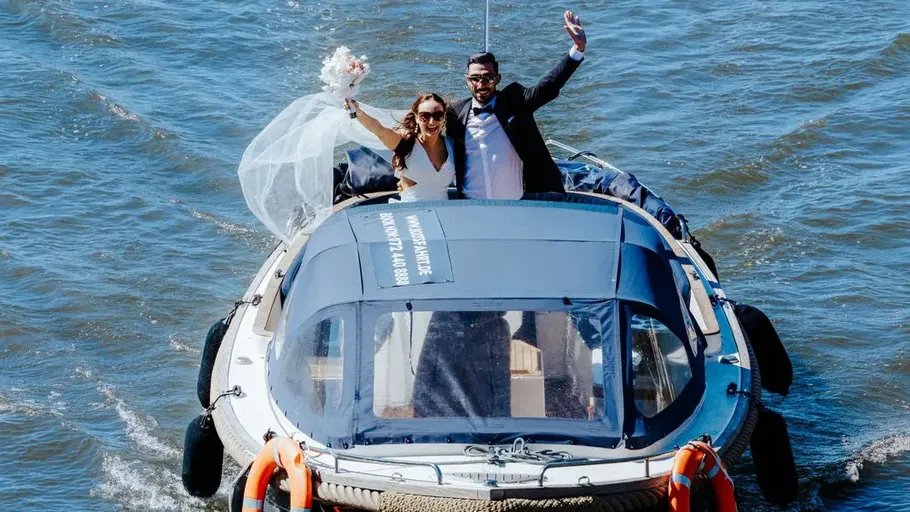 Brautpaar winkt auf Boot im Wasser.