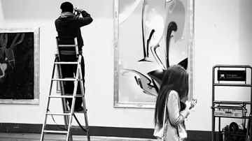 Man on ladder photographing street art, woman walking.