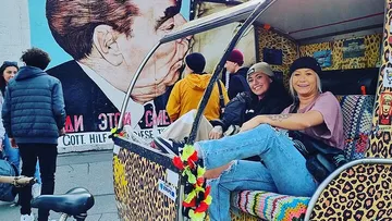 Two women sitting in colorful vehicle, mural behind.