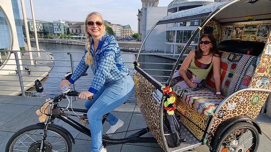 Woman cycling rickshaw, another woman sitting, riverside.