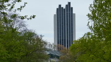 Hohes Gebäude hinter Bäumen und Gewächshaus.