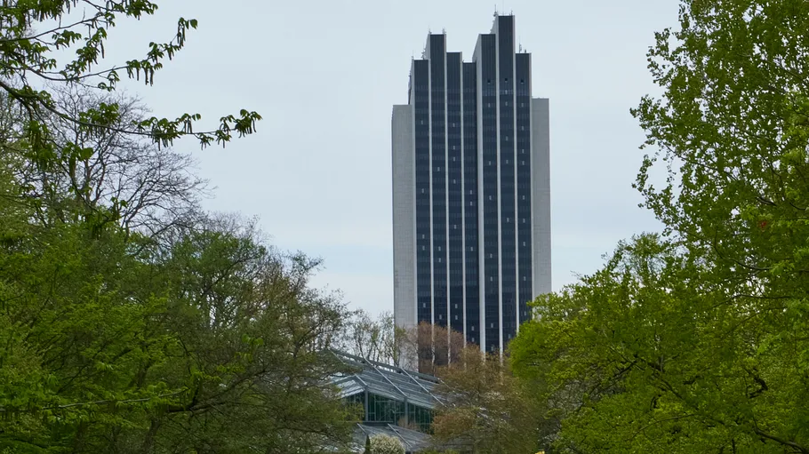 Hohes Gebäude hinter Bäumen und Gewächshaus.