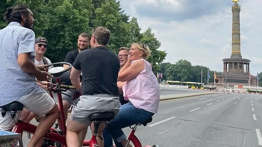 Gruppe radelt auf großem roten Fahrrad nahe Denkmal.