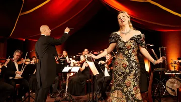 Woman singing with orchestra under red drapes.