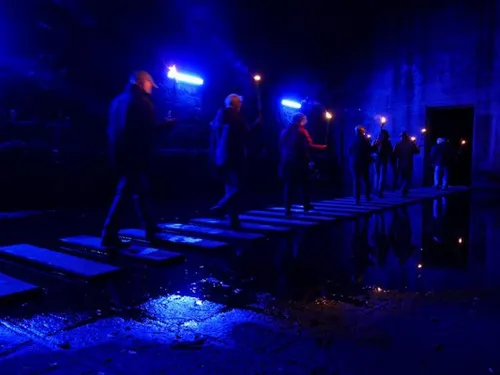 People walk on wooden planks across a dark, reflective waterway.