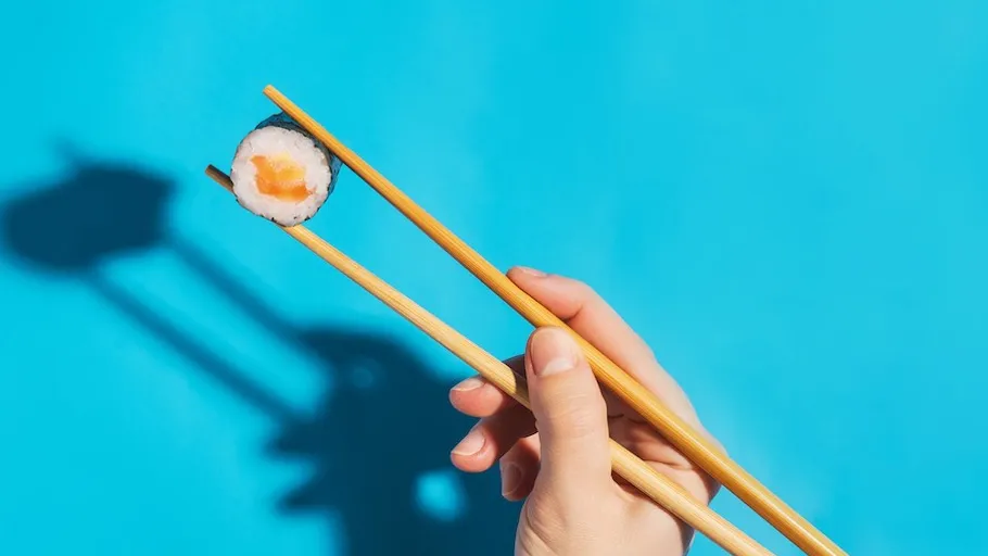 Hand hält Sushi mit Stäbchen, blauer Hintergrund.