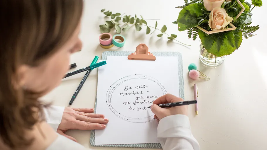 Person writing on paper, surrounded by pens, flowers.