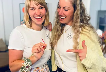 Two women smiling, holding floral wreath indoors.
