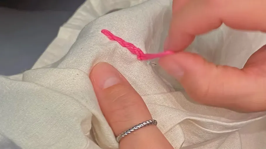 Hands embroidering pink thread on fabric.