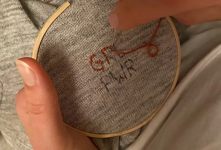 Hands embroidering text on gray fabric.