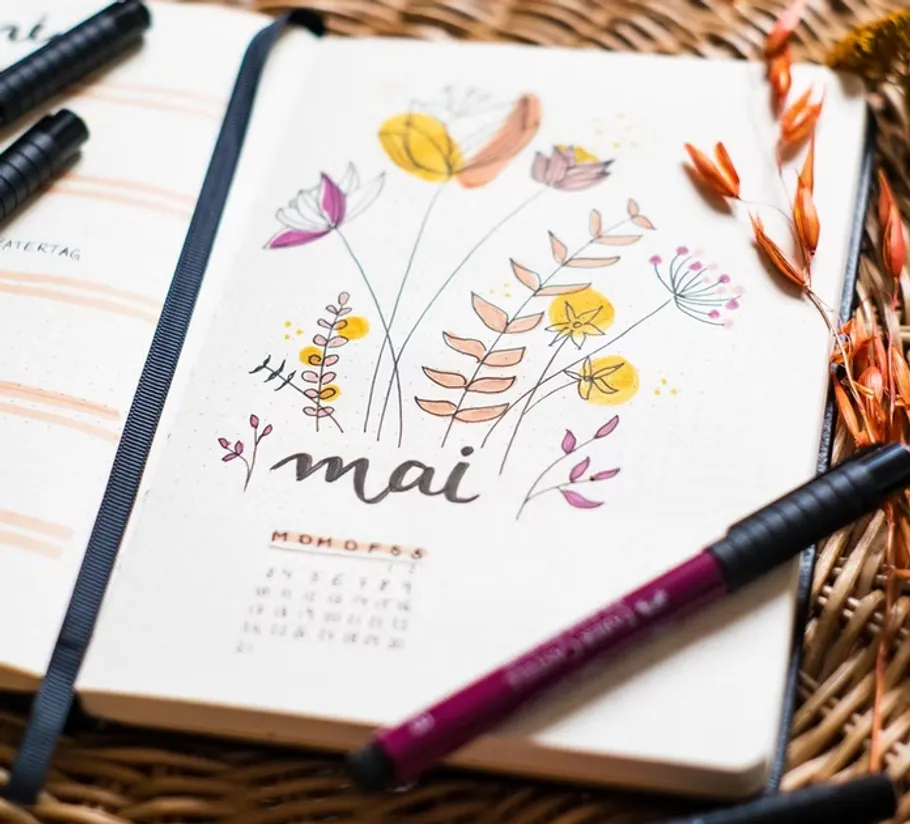 Notizbuch mit Mai-Blumenillustration, Stifte daneben.