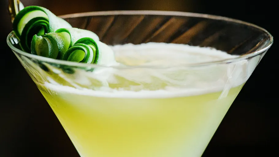 Green cocktail in glass with cucumber garnish.