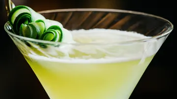 Green cocktail with cucumber garnish in glass.