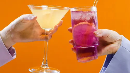 Two hands hold cocktails against orange background.
