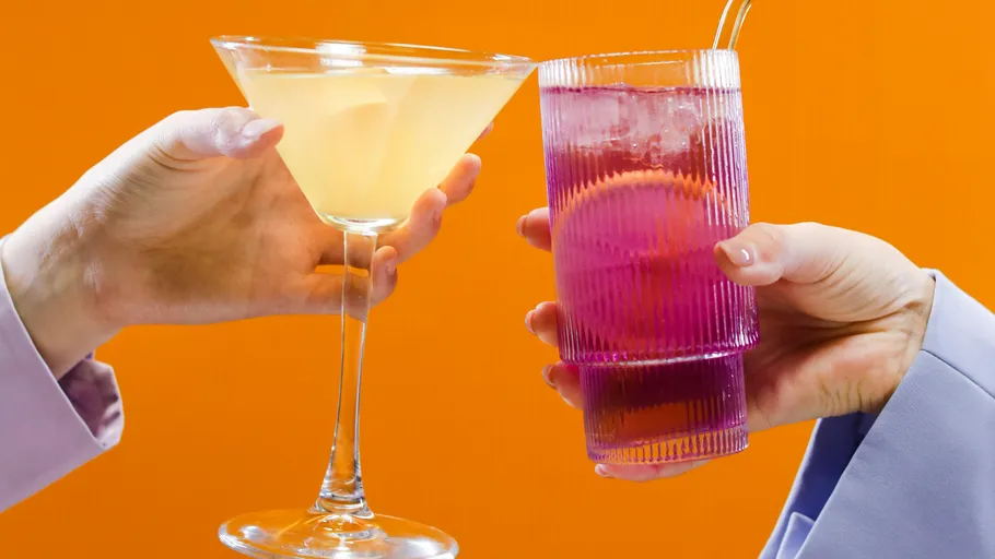 Zwei Hände halten Cocktails vor orangefarbenem Hintergrund.