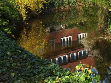 Water reflects a brick building surrounded by plants.
