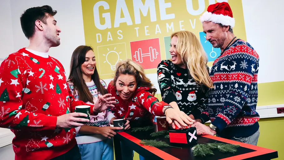 Gruppe lacht in Weihnachtspullovern am Spieltisch.