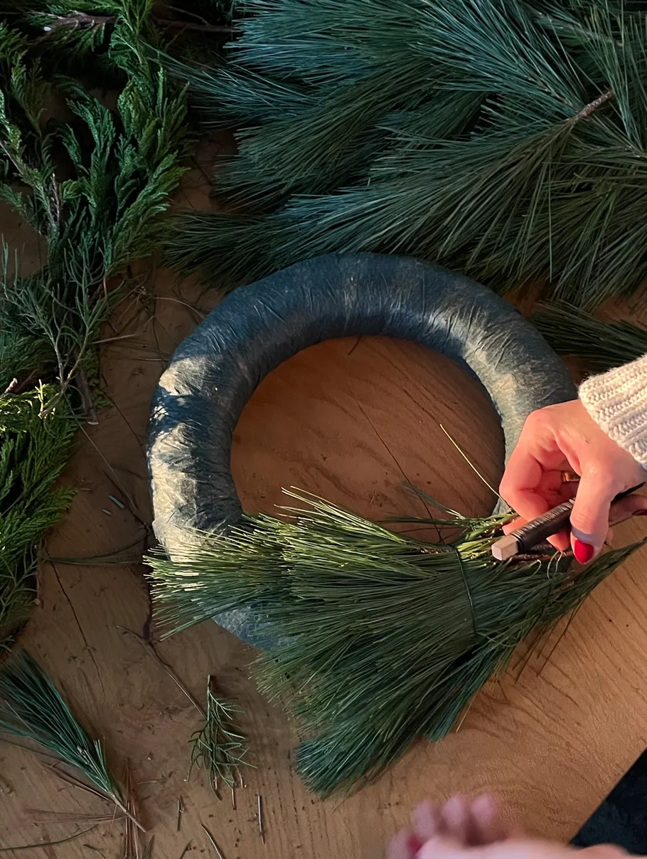 Person crafting wreath with pine branches indoors.