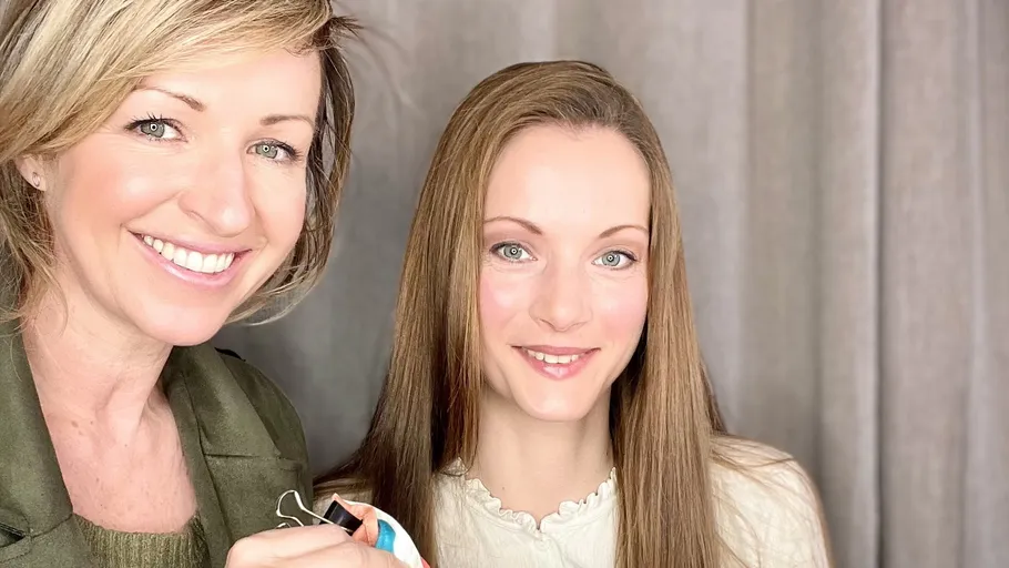Two smiling women indoors with a gray curtain.