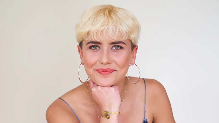 Person mit kurzem blondem Haar lächelt drinnen.
