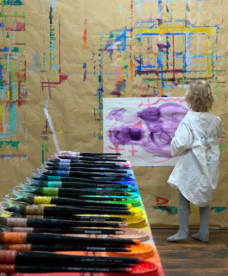 Child painting on colorful wall, art supplies nearby.