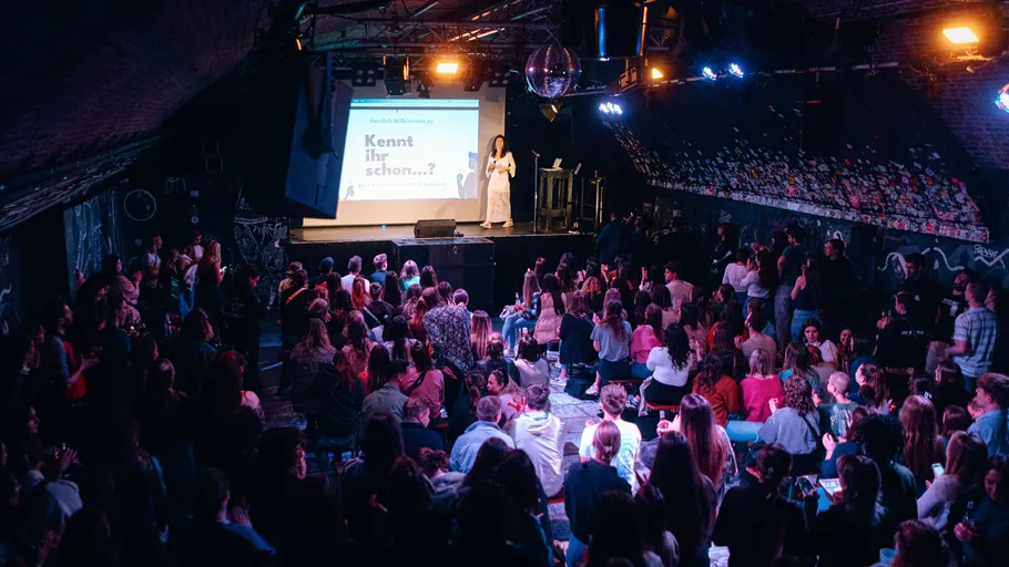 Crowded venue, speaker on stage, audience listening.