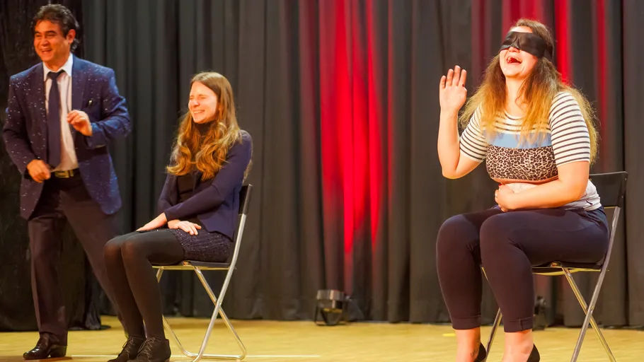 Blindfolded woman waves, seated on stage.