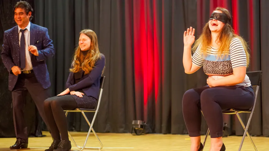 Three people on stage, two seated, one blindfolded.