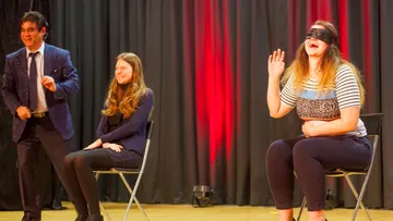 Two seated women, one blindfolded, man standing nearby.