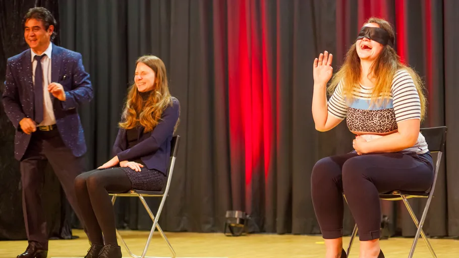 Three people on stage, one blindfolded, two seated.